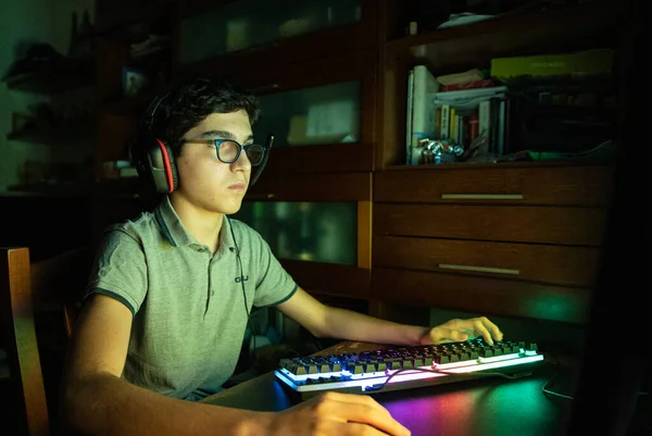 Game action during a video gaming session. The Caucasian boy with wavy black hair and glasses has his face illuminated by the changing colors of the screen during the game. Wear gaming headphones.