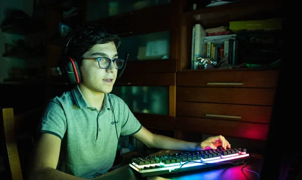 Game action during a video gaming session. The Caucasian boy with wavy black hair and glasses has his face illuminated by the changing colors of the screen during the game. Wear gaming headphones.