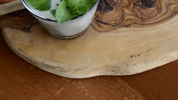 いくつかのミントの葉で豊かに白いケフィアの完全なガラスの上にクローズアップショット まな板にはケフィアの発酵が詰まった木製のスプーン — ストック動画
