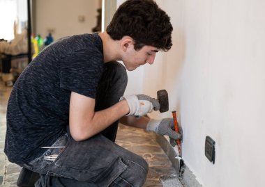 Apartmanda mason işi: beyaz bir çocuk çalışma eldiveni giyiyor ve eski süpürgeliği yıkmak için çekiç ve keski kullanıyor..