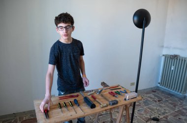 Beyaz bir çocuk masaya iş aletleri sipariş etmek niyetindedir: tornavida, çekiç, pense, alet bıçağı ve koruyucu gözlük.