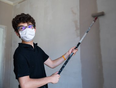Koronavirüs zamanında evin duvarlarını boyayarak onları yenile: Beyaz çocuğun kendini virüsten korumak için bir maskesi var. Hamile bırakılmış gri renk silindirini duvarda gezdir..