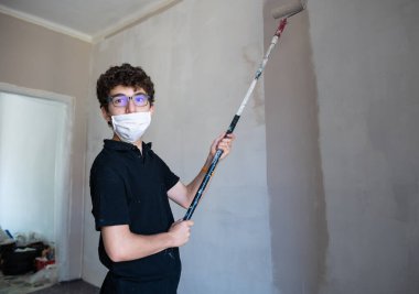 Koronavirüs zamanında evin duvarlarını boyayarak onları yenile: Beyaz çocuğun kendini virüsten korumak için bir maskesi var. Hamile bırakılmış gri renk silindirini duvarda gezdir..