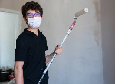 Koronavirüs zamanında evin duvarlarını boyayarak onları yenile: Beyaz çocuğun kendini virüsten korumak için bir maskesi var. Hamile bırakılmış gri renk silindirini duvarda gezdir..