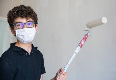 Koronavirüs zamanında evin duvarlarını boyayarak onları yenile: Beyaz çocuğun kendini virüsten korumak için bir maskesi var. Hamile bırakılmış gri renk silindirini duvarda gezdir..