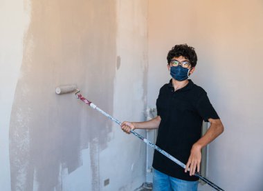 Koronavirüs zamanında evin duvarlarını boyayarak onları yenile: Beyaz çocuğun kendini virüsten korumak için bir maskesi var. Hamile bırakılmış gri renk silindirini duvarda gezdir..