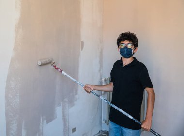 Koronavirüs zamanında evin duvarlarını boyayarak onları yenile: Beyaz çocuğun kendini virüsten korumak için bir maskesi var. Hamile bırakılmış gri renk silindirini duvarda gezdir..