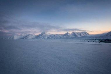 Pembe ve mavi tonlarda yumuşak karlı yüzey ve arka planda dağlar.