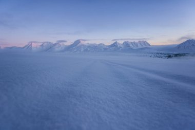 Pembe ve mavi tonlarda yumuşak karlı yüzey ve arka planda dağlar.