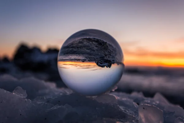 Buzlu kar üzerindeki cam bir kürenin fotoğrafı. İçinde manzaranın ters yansıması var.