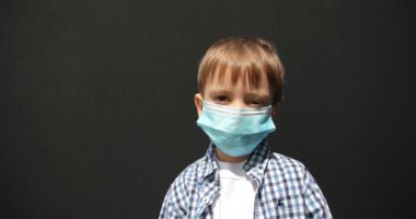 Koronavirüs salgını sırasında gri duvara bakarken tıbbi maskeli çocuk kameraya bakıyor. Tıbbi maskeli küçük bir çocuk gri bir arka planda duruyor..