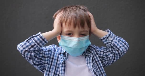 Un niño pequeño con una máscara médica en su cara sostiene su cabeza en sus manos de miedo — Vídeos de Stock