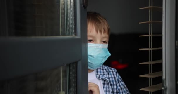 Porträt eines kleinen Jungen mit medizinischer Gesichtsmaske, der am Fenster steht. Trauriges männliches Kind mit Schutzmaske vor dem Virus draußen. Konzept der Gesundheit und Sicherheit vor Coronavirus und Pandemie. — Stockvideo