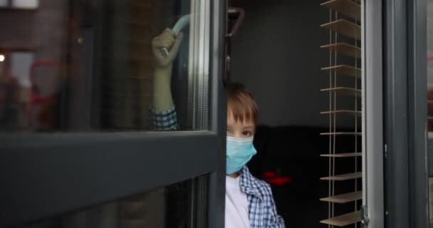 Ένα μικρό αγόρι με μάσκα ανοίγει παράθυρο ή πόρτα. Χαριτωμένο μικρό παιδί που φοράει ιατρική μάσκα κατά τη διάρκεια της επιδημίας covid-19 ή coronavirus. — Αρχείο Βίντεο