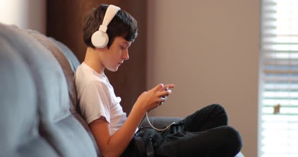 Um adolescente senta-se em um sofá com fones de ouvido brancos e joga um jogo em um smartphone. Menino jogando jogos no smartphone sentado em casa durante a quarentena . — Vídeo de Stock