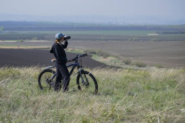 Bisikletli bir kadın tepede durup su içiyor..