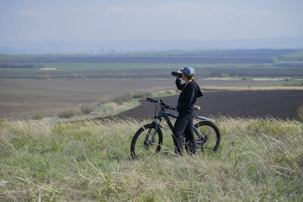 A woman on a bike stopped on a hill and drinks water.