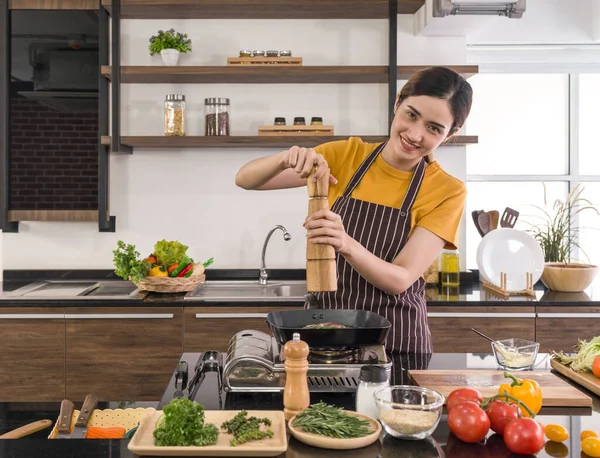 Young housewife putting pepper on the steak. Morning atmosphere in a modern kitchen. The kitchen counter full of various kinds of vegetables and seasonings.