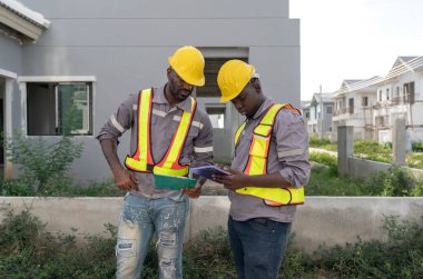 İnşaat alanındaki tablette miğfer ve güvenlik yeleği takan iki inşaat işçisi. Proje detaylarını tartışırken dikkatle görevlerine odaklanırlar.