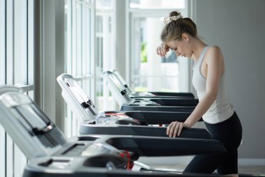 Koşu bandında egzersiz yapan bir kadın, geniş pencereli, parlak, çağdaş bir spor salonunda soluklanır..