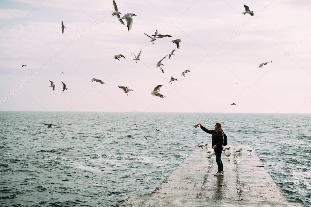  A girl stands near the sea on a pier with a backpack and feeds the seagulls.