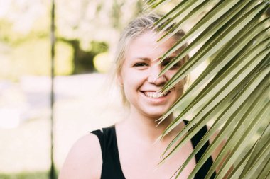  Genç bir kızın yüzünü kaplayan yeşil bir palmiye dalına yakın plan. Arka planda yeşil bokeh yaprakları olan siyah kolsuz tişörtlü bulanık bir kızın portresi. Sonbaharda palmiye ağaçlarının yanında dinlen.