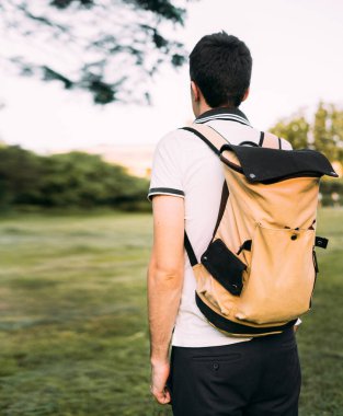 Omzunda bej ve siyah bir sırt çantası olan beyaz tişörtlü ve şortlu genç bir adam ayağa kalkar ve