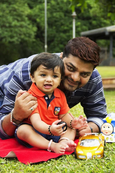 Padre e hijo pequeño, sonriendo mirando a la cámara con juguetes alrededor, mientras está sentado dentro del jardín, Pune, Maharashtra . — Foto de Stock