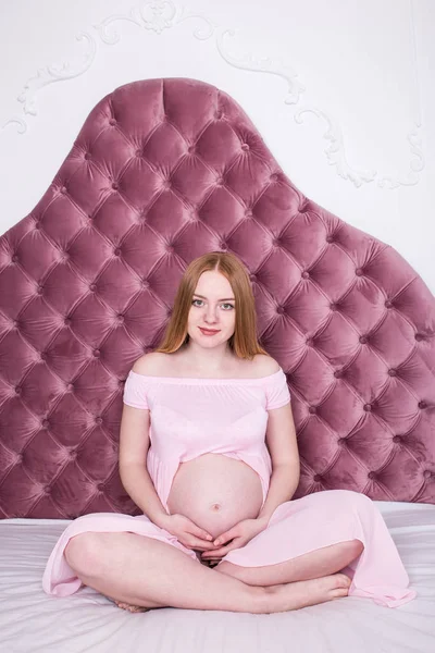 Gebelik, sevgi, insanlar ve beklenti kavramı. Pembe elbiseli yatağın üstüne oturan hamile kadın. 