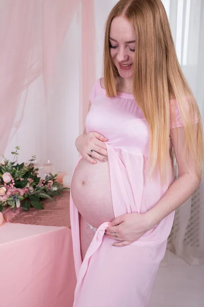 妇女在怀孕期间在粉红色礼服站立在窗口附近 — 图库照片