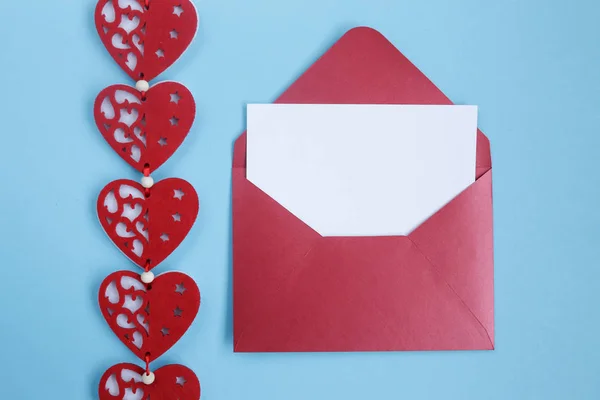 Open red envelope with blank white gift card and red hearts decoration on blue background. Valentines day background with hearts. Copyplace, space for text and logo.