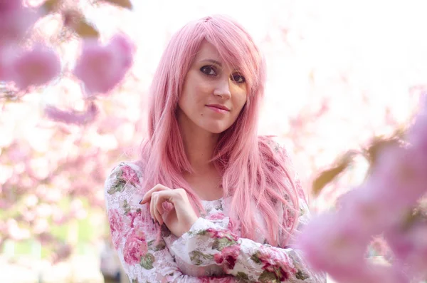 Jonge mooie vrouw met roze haar in de buurt van sakura bloeiende boom. — Stockfoto