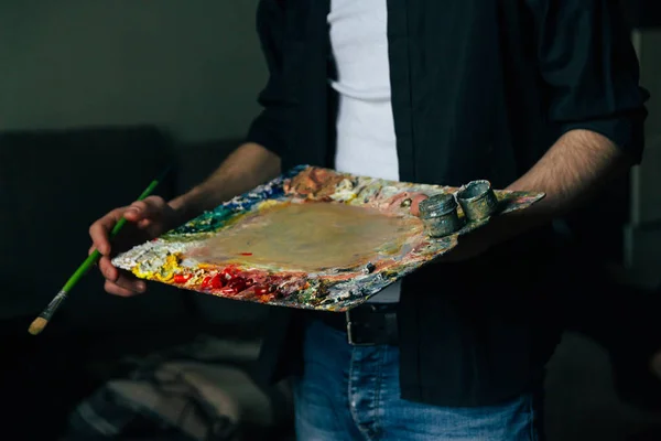 El artista sostiene una paleta con pinturas y un pincel. un hombre en jeans y una camisa negra . — Foto de Stock