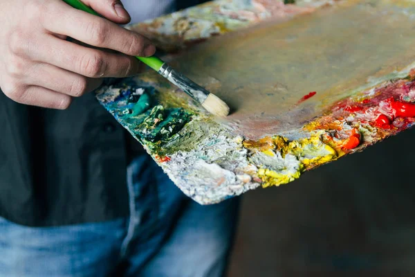 El artista sostiene una paleta con pinturas y un pincel y va a pintar sobre lienzo. Mezclar las pinturas al óleo — Foto de Stock
