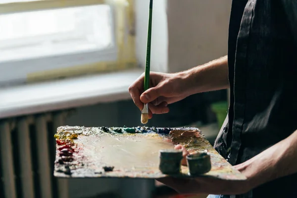 Frente a la ventana el artista sostiene una paleta con pinturas y un pincel y va a pintar sobre lienzo — Foto de Stock