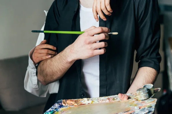 Un hombre sostiene en su mano una paleta con pinturas y un pincel, y detrás de él abrazan — Foto de Stock