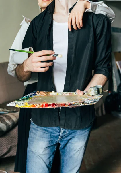 Un hombre sostiene en su mano una paleta con pinturas y un pincel, y detrás de él abrazan — Foto de Stock