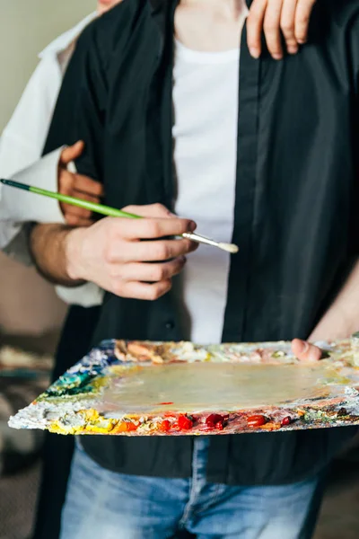 a man holds in his hand a palette with paints and a brush, and behind him they embrace