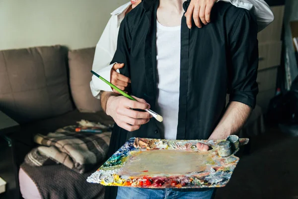 Un hombre sostiene en su mano una paleta con pinturas y un pincel, y detrás de él abrazan —  Fotos de Stock