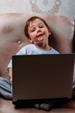 bir sandalye, kanepe bir dizüstü bilgisayar ile küçük çocuk beyaz bir tişört içinde oturur. fotoğraf makinesine benziyor. komik surat. konsantrasyon.