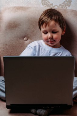 bir sandalye, kanepe bir dizüstü bilgisayar ile küçük çocuk beyaz bir tişört içinde oturur. fotoğraf makinesine benziyor. komik surat. konsantrasyon.