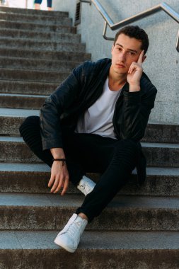 Seksi erkek poz korkuluk yakınındaki adımları oturur. Yakışıklı delikanlı şık siyah elbise ve Beyaz Ayakkabı yakınındaki bir korkuluk poz