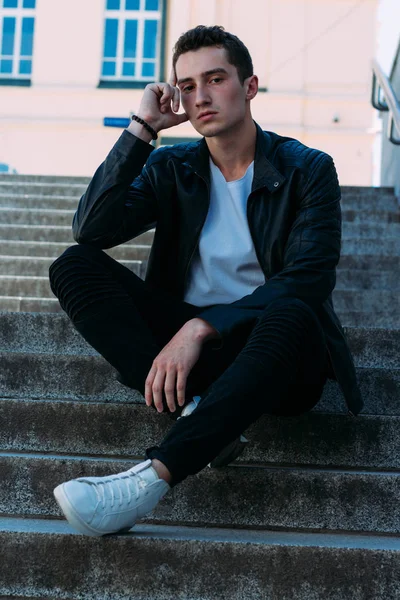 Hombre sexy posando se sienta en las escaleras cerca de la barandilla. Guapo joven en ropa elegante negro y zapatos blancos posando cerca de una barandilla — Foto de Stock