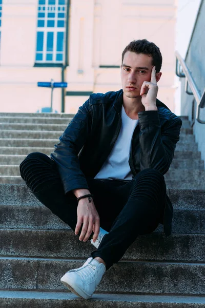 Sexy Mann posiert auf den Stufen neben Geländer. schöner junger Mann in stylischer schwarzer Kleidung und weißen Schuhen, der an einem Geländer posiert — Stockfoto