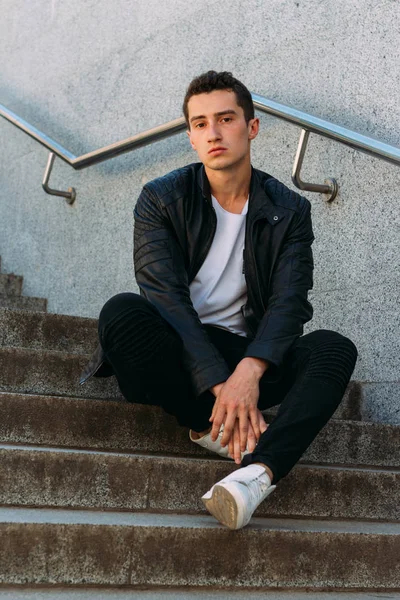Seksi erkek poz korkuluk yakınındaki adımları oturur. Yakışıklı delikanlı şık siyah elbise ve Beyaz Ayakkabı yakınındaki bir korkuluk poz