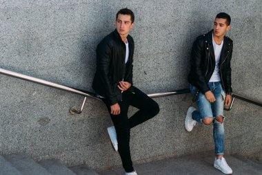 beklenti seksi poz korkuluk yakınındaki mans. çekici bir genç adamın bir siyah deri ceket, beyaz bir T-shirt ve beyaz spor ayakkabı korkuluk yakın duruyor. dostluk. iki model. Giysi gösterilen