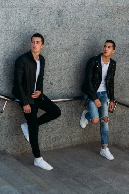 beklenti seksi poz korkuluk yakınındaki mans. çekici bir genç adamın bir siyah deri ceket, beyaz bir T-shirt ve beyaz spor ayakkabı korkuluk yakın duruyor. dostluk. iki model. Giysi gösterilen