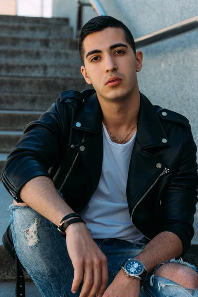 Seksi erkek poz korkuluk yakınındaki adımları oturur. çekici bir genç adam bir siyah deri ceket, beyaz bir gömlek ve beyaz spor ayakkabı. yandan izle