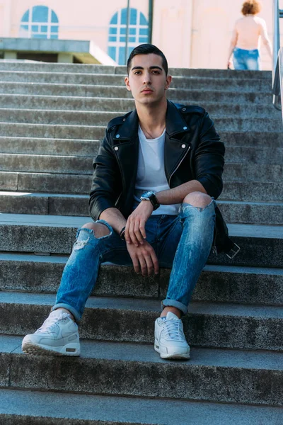 Seksi erkek poz korkuluk yakınındaki adımları oturur. çekici bir genç adam bir siyah deri ceket, beyaz bir gömlek ve beyaz spor ayakkabı. yandan izle