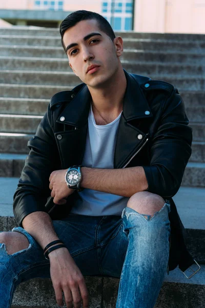 Seksi erkek poz korkuluk yakınındaki adımları oturur. çekici bir genç adam bir siyah deri ceket, beyaz bir gömlek ve beyaz spor ayakkabı. yandan izle